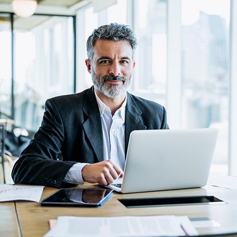 Smiling bearded businessman working on laptop in office Buenos Aires businessman sitting at office work table next to window with view of downtown and pausing from typing on laptop to smile at camera.