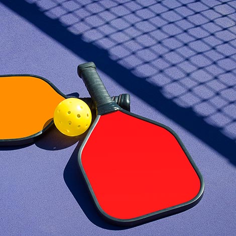 Imae of 2 Pickleball paddles and a pickleball laying on pickleball court with net shadow.