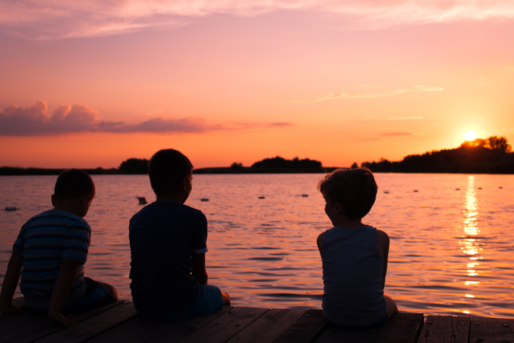 Three boys sitting on pier by sea at sunset. Relaxed kids enjoying in sunset at beach.