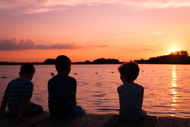 Rear view of kids on pier at sunset. Three boys sitting on pier by sea at sunset. Relaxed kids enjoying in sunset at beach.