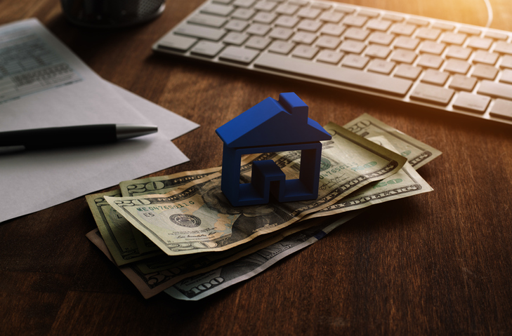 3D house on pile of money with tax and financial forms on the desk.