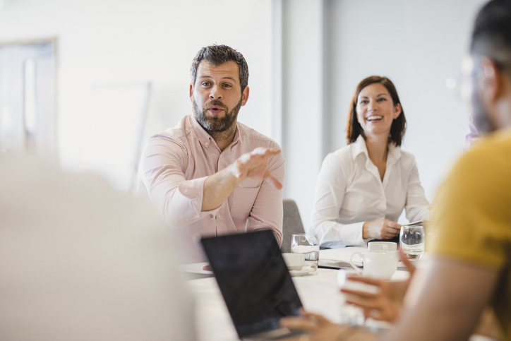 Mature man in his 40s gesturing and talking, sitting at meeting table in board room, discussion, collaboration, strategy