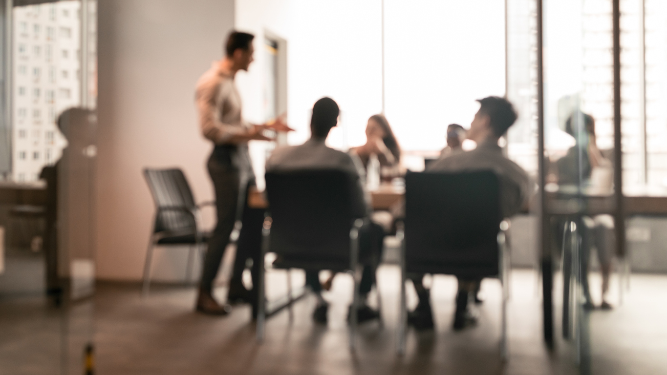 Colleagues having meeting in boardroom, businessman giving speech, blurred photo Business Presentation, Blurred Background. Businessman Giving Speech During Seminar With Coworkers In Office, Standing At Desk In Boardroom, Diverse People Sitting At Table And Listening To Speaker