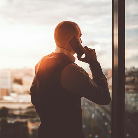 Silhouette of a prosperous man entrepreneur standing near the panoramic window of a luxurious business office skyscraper and pensively looking on an evening cityscape while talking on the phone