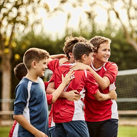 Happy soccer players are huddling on ground. Boys are celebrating success after match. They are on sports field.