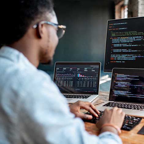 Young african male programmer writing program code sitting at the workplace with three monitors in the office. Image focused on the screen