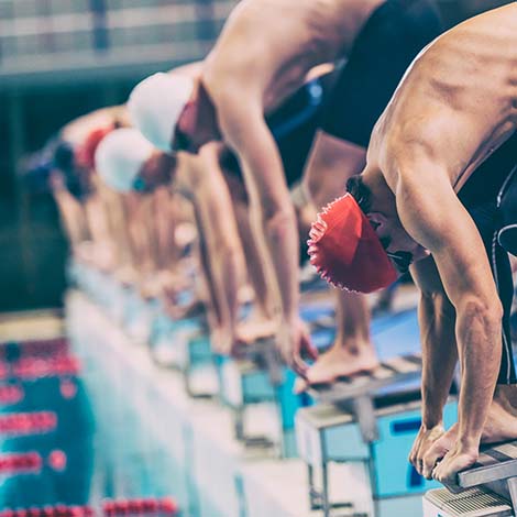 Close up shot of a male swimmer crouching on starting blocks ready to jump into the water at the signal, other swimmers defocused.