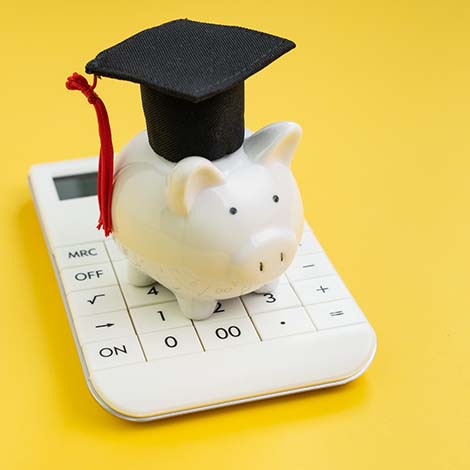 Student loan payment calculation, scholarship or saving for school and education concept, white piggy bank wearing graduation hat on calculator on yellow background with copy space Student loan payment calculation, scholarship or saving for school and education concept, white piggy bank wearing graduation hat on calculator on yellow background with copy space.