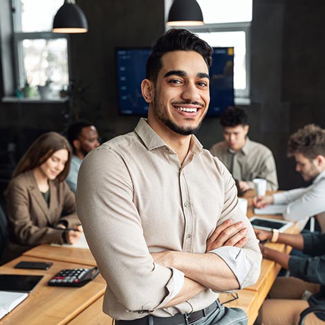 Successful Person. Portrait of confident smiling bearded businessman sitting leaning on desk in office, posing with folded arms and looking at camera, colleagues working in blurred background