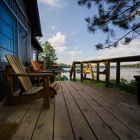 Two Muskoka chairs on a wooden deck facing a lake. In the background there’s a pier with a big amount of chairs