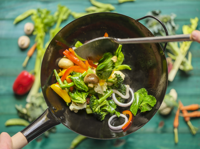 A variety of fresh market vegetables including broccoli, carrot, capsicum, snow peas, red onion, green beans, celery and mushrooms being stir-fried and cooked in a hot wok, with an out of focus turquoise-colored wooden, vegetable covered table in the background.