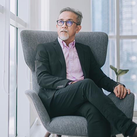 Successful Asian chinese senior businessman in suit sitting on sofa in his office room looking at the capital city