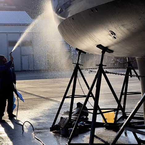 Blue collar worker spraying water on the bottom of private boat. Beautiful sun light reflecting on water drops. Yacht lifted for maintenance and painting works on dry dock. Professional occupation and service at shipyards.