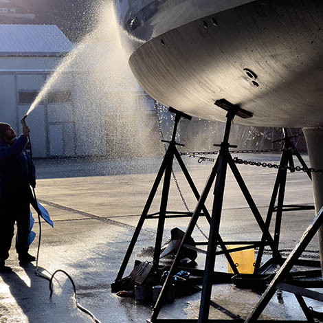 Man silhouette washing the boat hull, side view Blue collar worker spraying water on the bottom of private boat. Beautiful sun light reflecting on water drops. Yacht lifted for maintenance and painting works on dry dock. Professional occupation and service at shipyards.