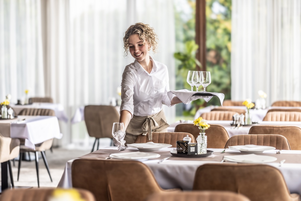 A smiling waitress sets a beautifully arranged table in a bright and elegant restaurant. She neatly prepares the table with a white tablecloth, plates, folded napkins, wine glasses and cutlery. The interior is large, luxurious and welcoming