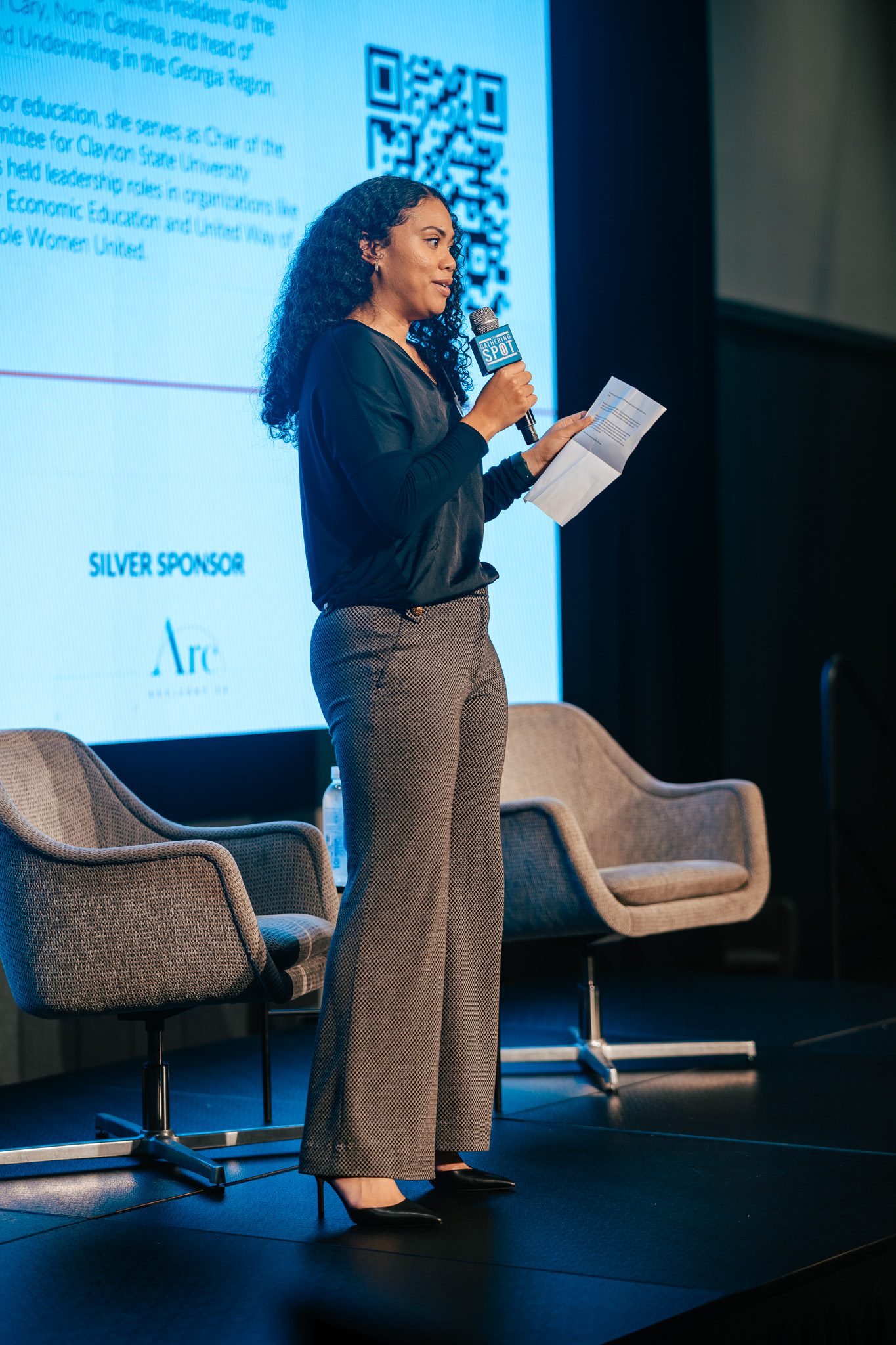 A woman stands on stage holding a microphone and papers, speaking in front of a screen with text and a QR code, next to two empty chairs.