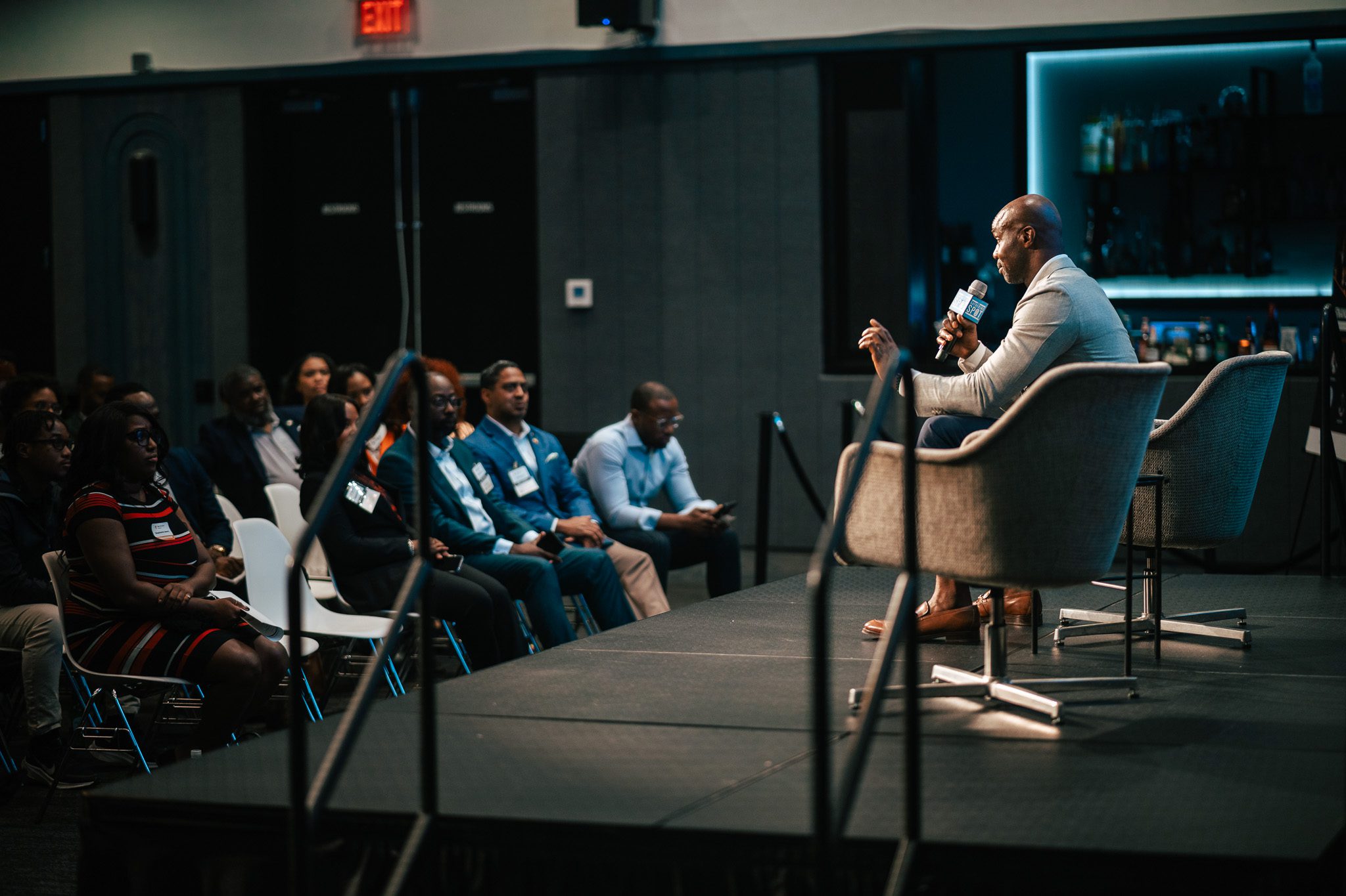 A man speaks into a microphone while seated on stage in front of an audience at an indoor event.