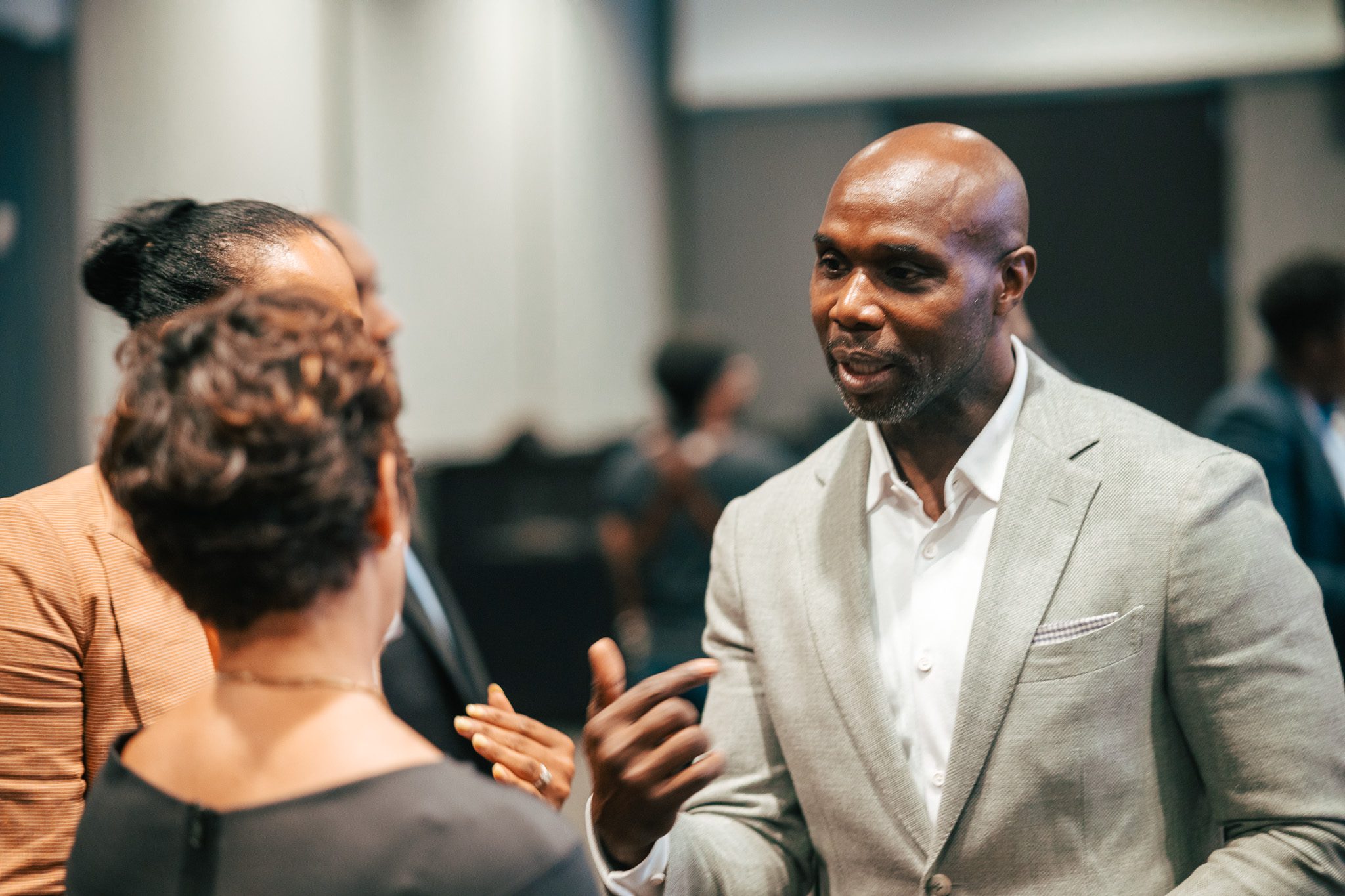 Three adults in business attire engage in conversation at an indoor event. The man in a light gray suit gestures while speaking to two women in the foreground.