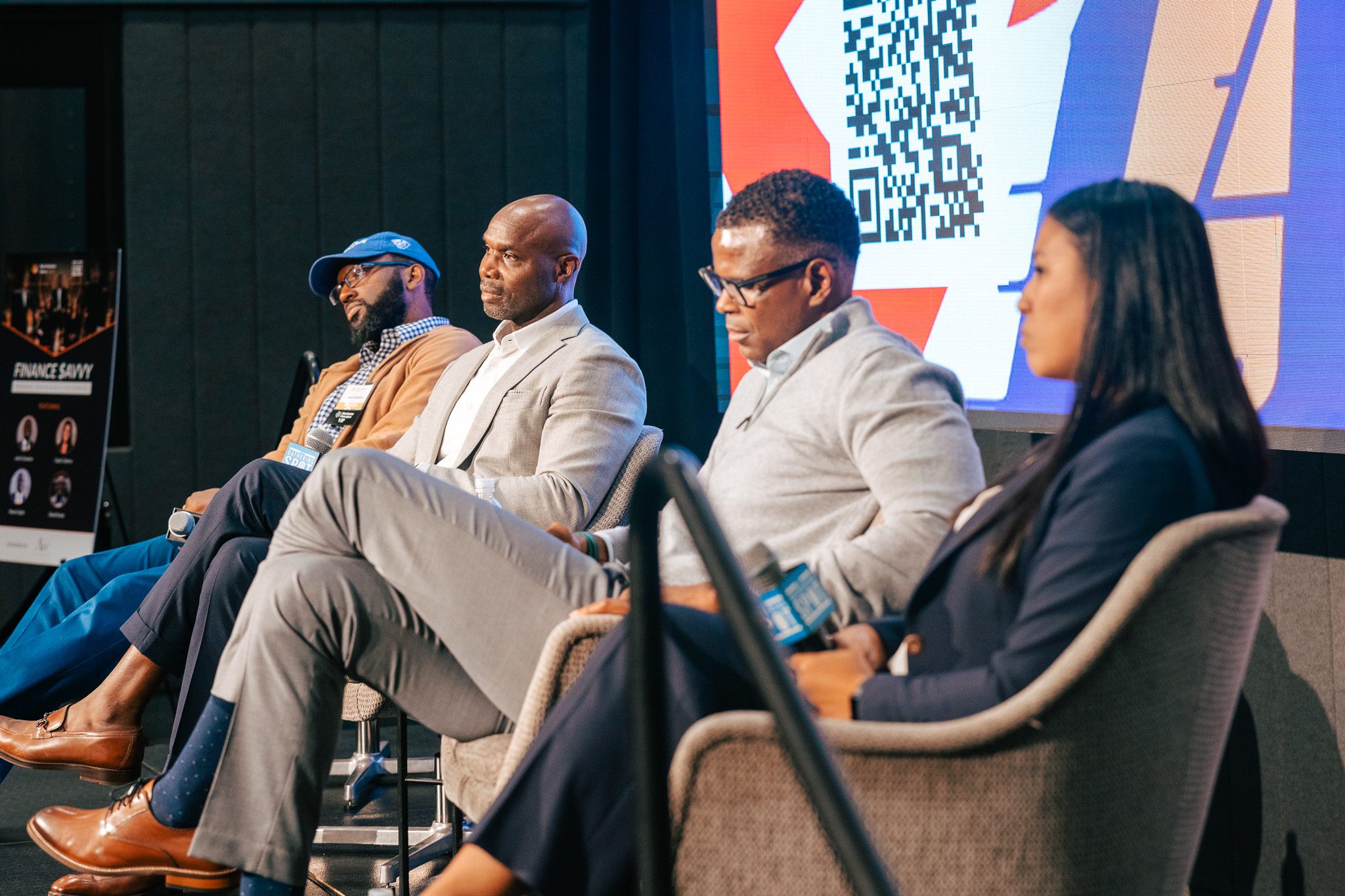 Four people sit in chairs on a stage, participating in a panel discussion; a large digital screen with a QR code is visible in the background.