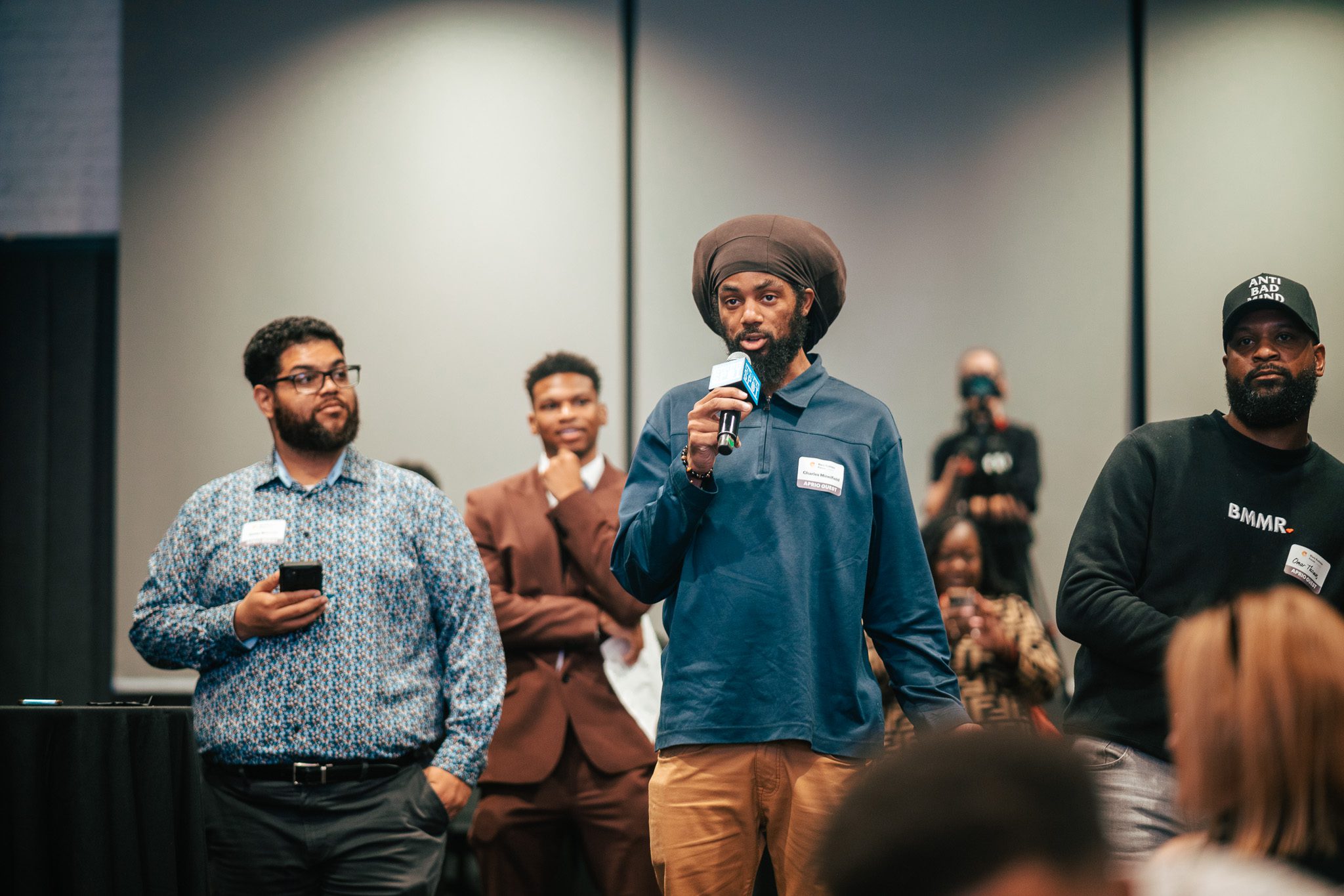 A man in a brown headwrap speaks into a microphone while others stand nearby and an audience watches in a conference room.