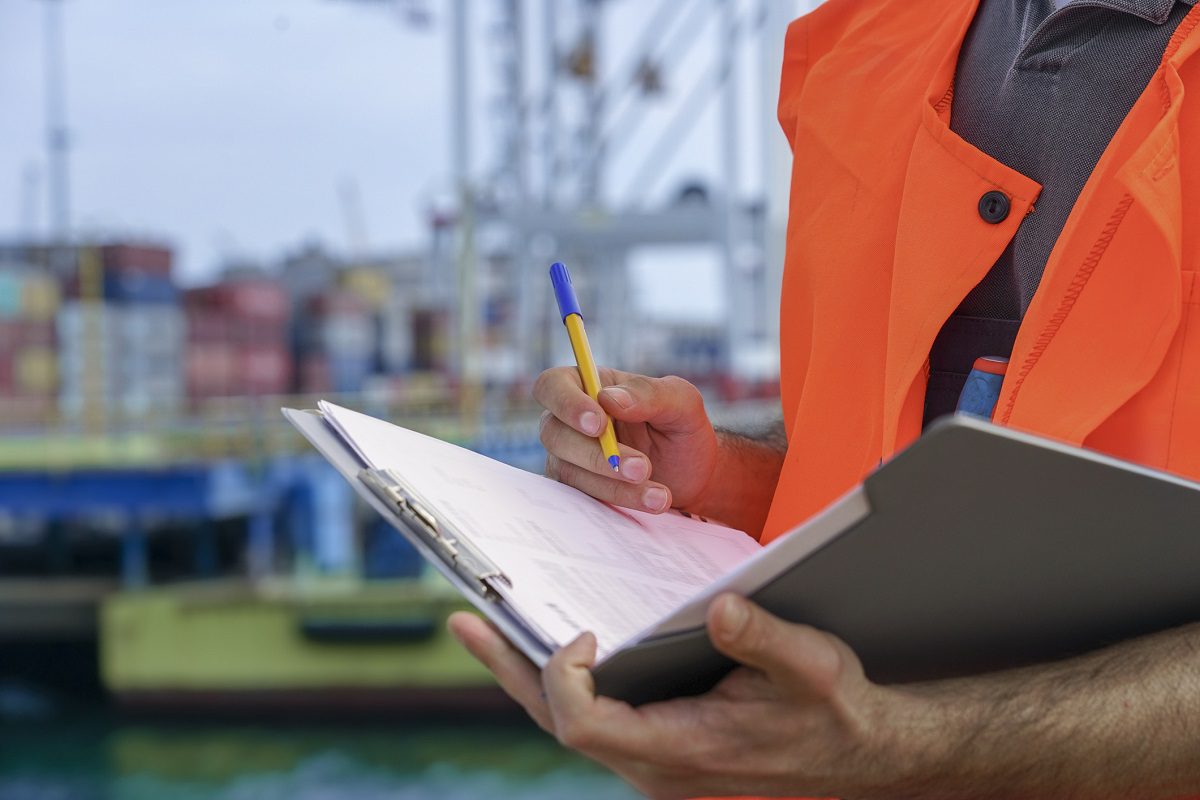 Close up of a man in an orange reflective shirt checking a clipboard in front of a blurred seaport background