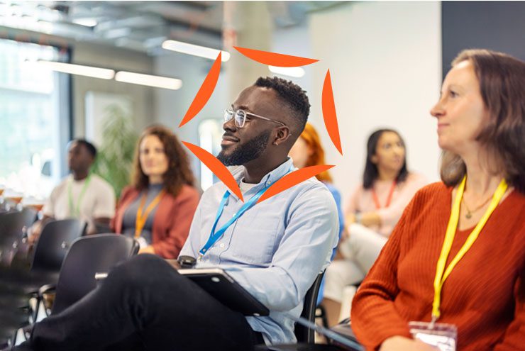 A man in glasses sits in a crowd and looks at something off camera. An Aprio logo has been superimposed around his face.
