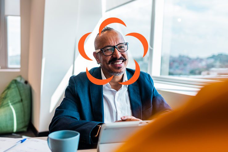 Man with glasses smiles at someone off camera. An Aprio logo has been superimposed around his face.