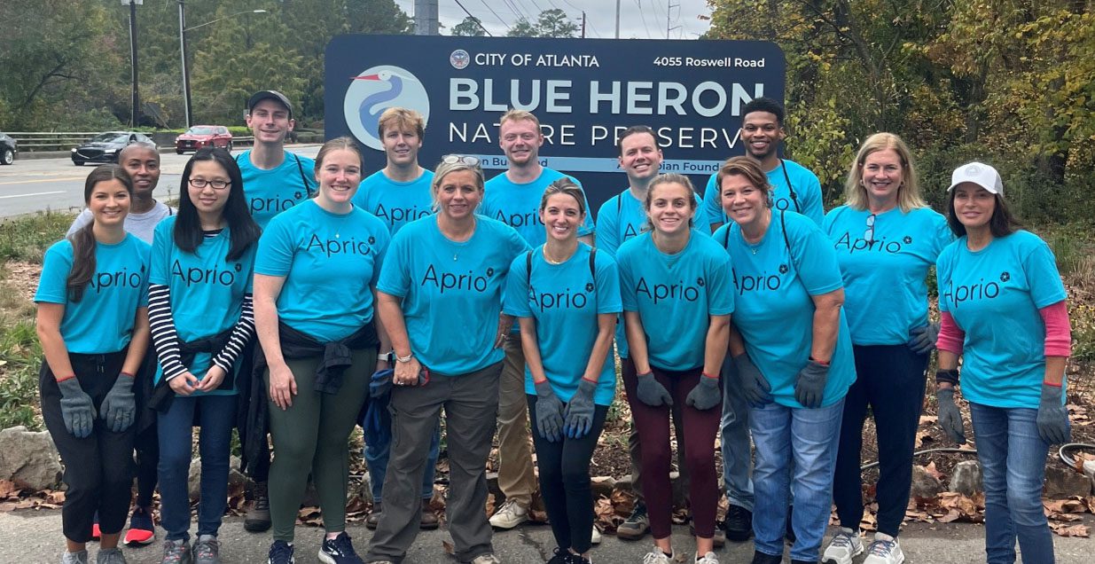 A group of volunteers in matching blue Aprio tshirts smile for the camera in front of a sign for the Blue Haron Nature Preserve
