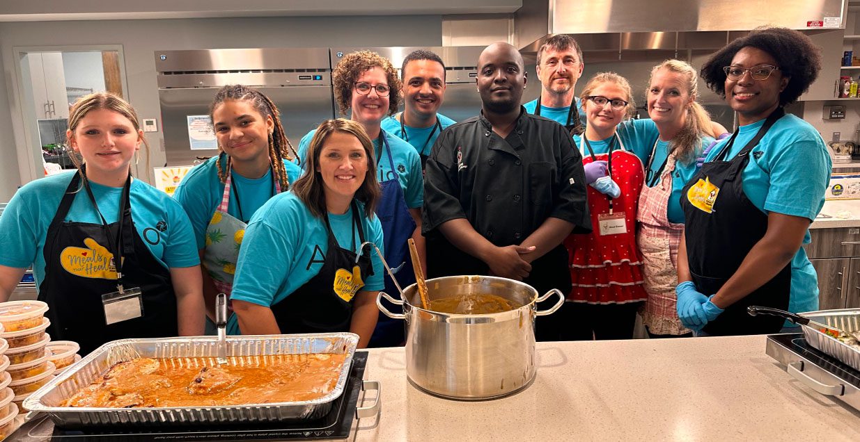 A group of Aprio volunteers in matching blue tshirts stand with a chef in a kitchen and smile for the camera
