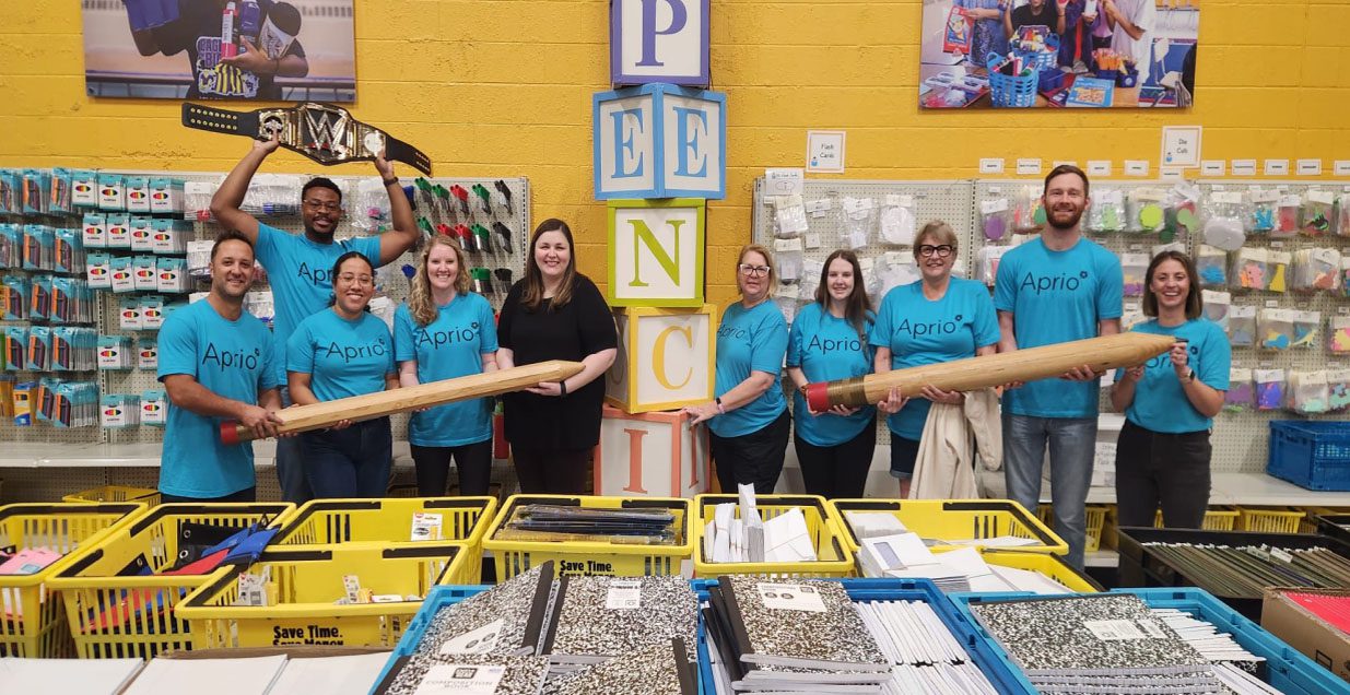 Volunteers in blue Aprio tshirts stand in a school supplies store holding giant pencils