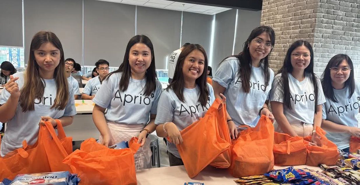 Volunteers in light blue Aprio tshirts fill tote bags with supplies and smile for the camera