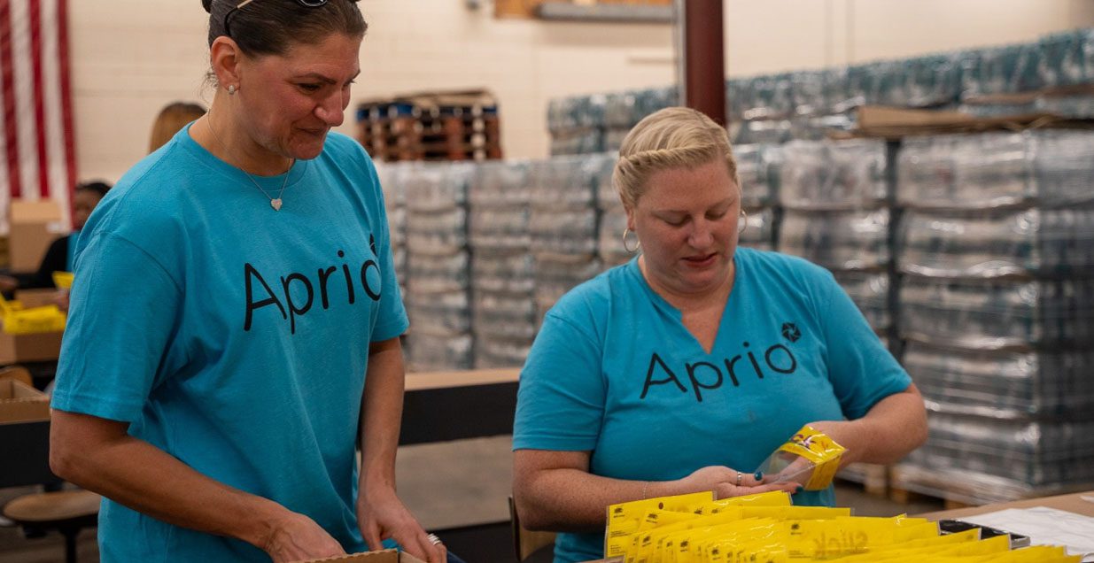 Two women in matching blue Aprio tshirts package supplies in a warehouse