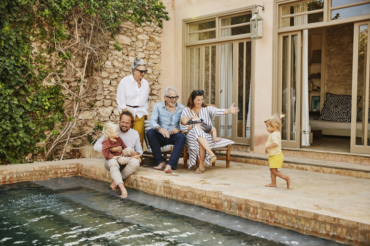 Wide shot of smiling grandparents relaxing with children and grandchildren beside pool at tropical villa during multigenerational family vacation