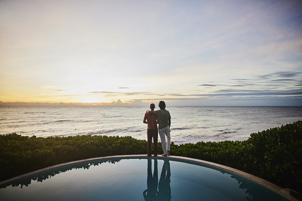 Wide shot of embracing couple watching sunrise while standing at edge of pool at luxury tropical villa overlooking ocean