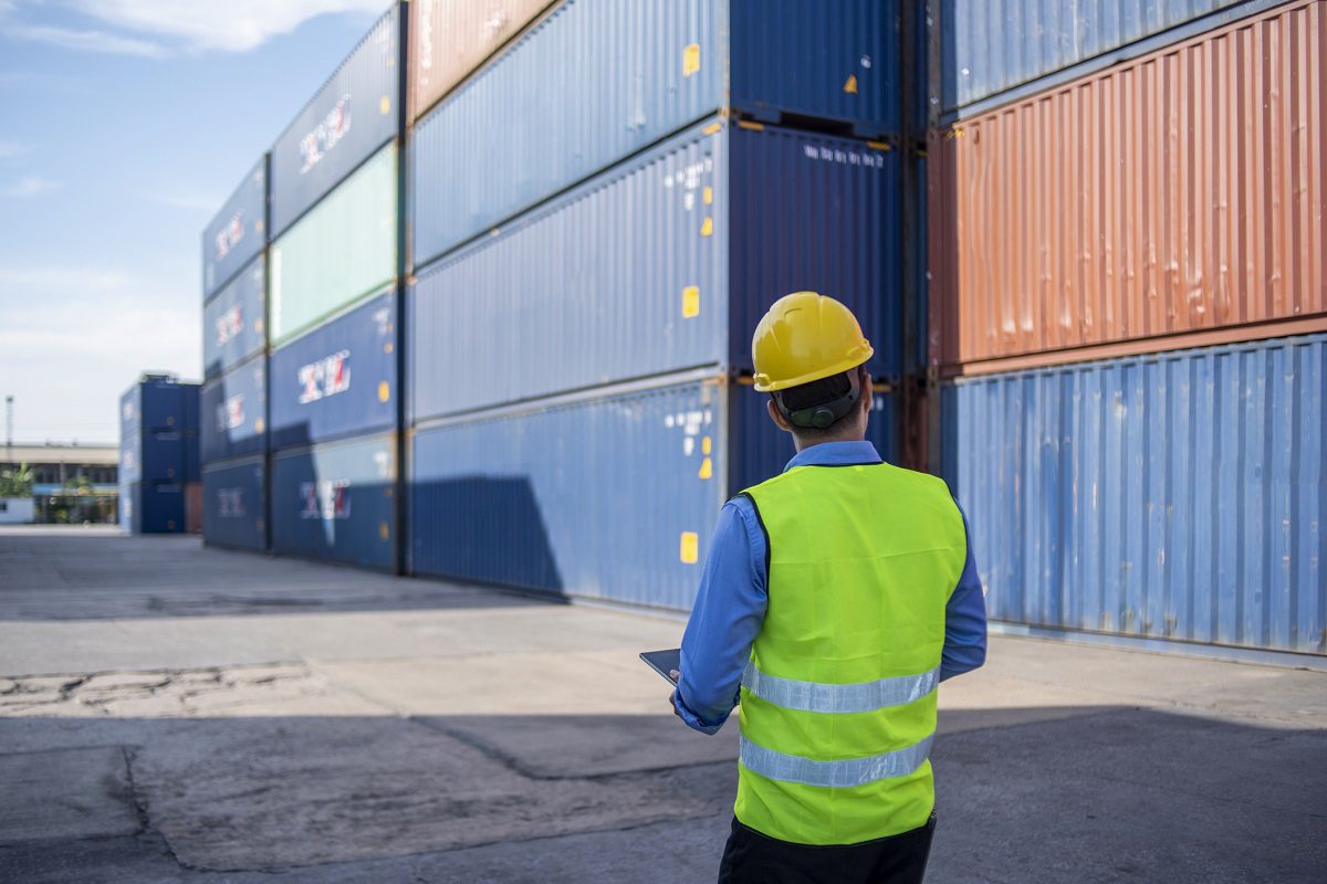 Shot from behind, a man in a hard hat and reflective vest walks through a row of stacked cargo containers in a shipyard