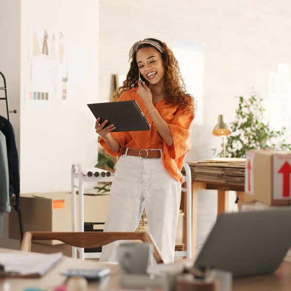 Young woman stands in a bright, half-unpacked office, looks at a tablet, and smiles while on her cellphone