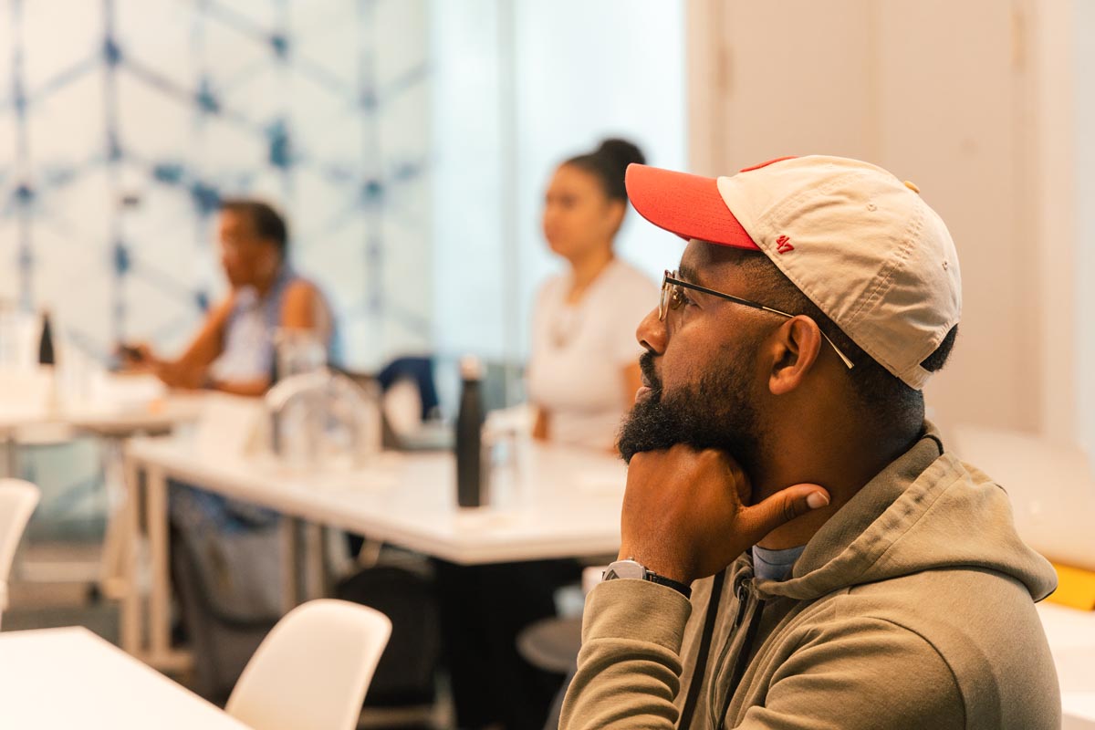 A man wearing a cap and glasses sits at a desk in a classroom, listening attentively. Two other people are seated at desks in the background.