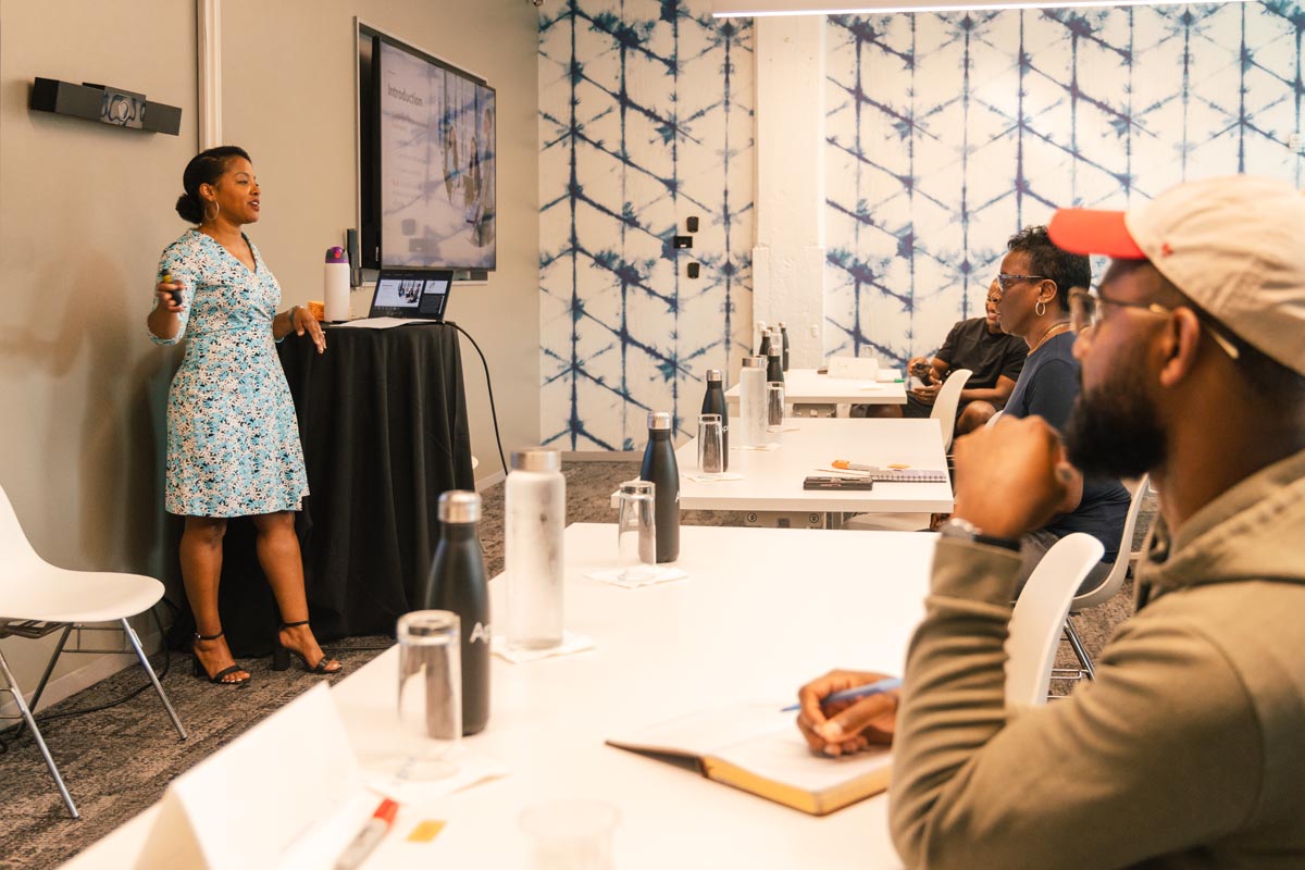 A woman stands and presents to a seated group in a conference room, pointing at a screen displaying a presentation.
