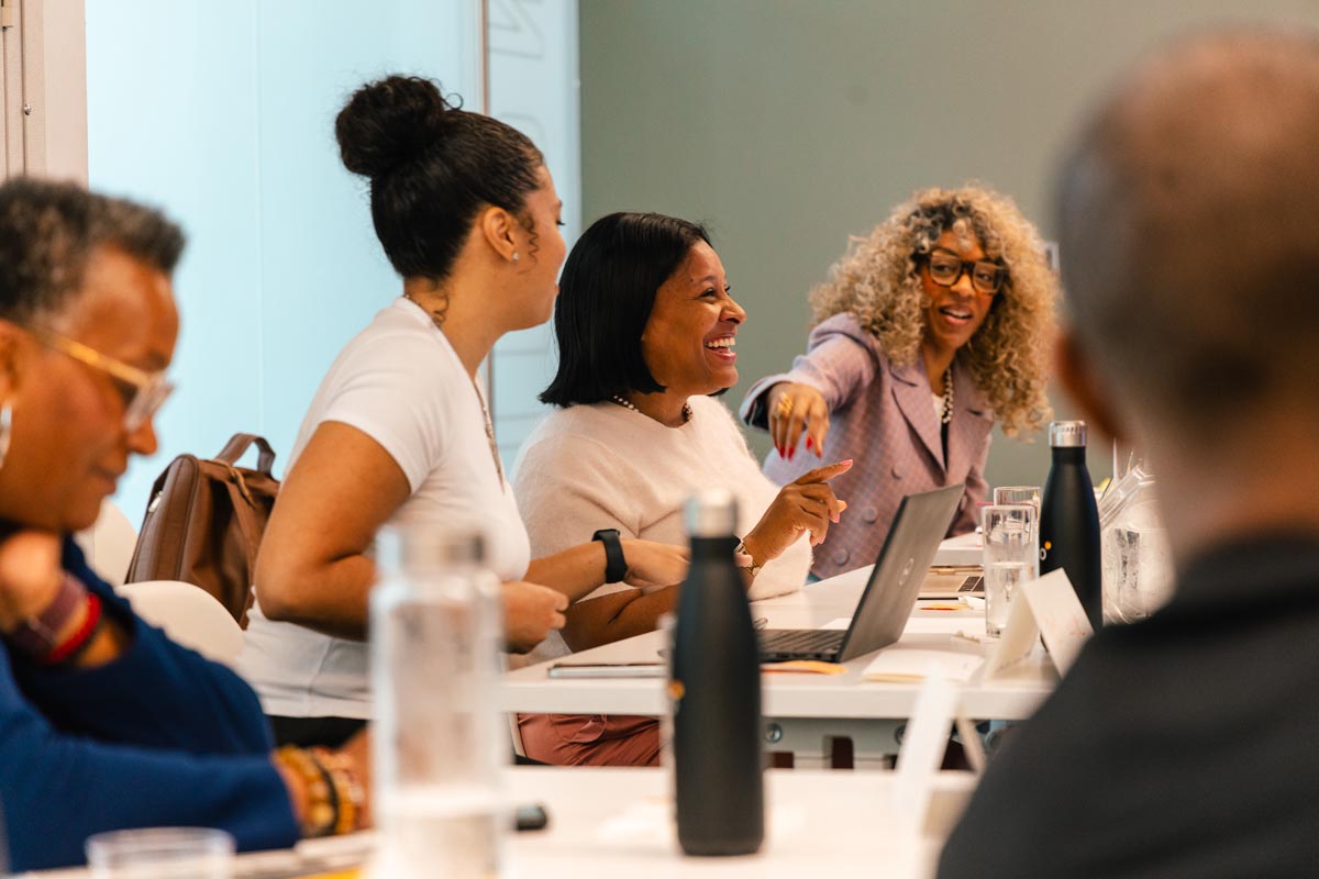 Four people sit at a conference table engaged in discussion, with papers, laptops, and water bottles in front of them. One person is smiling and pointing while others listen and talk.