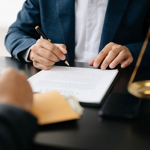 Shot from the chest down, a man in a suit jacket sits at a table and hovers a pen over a stack of documents