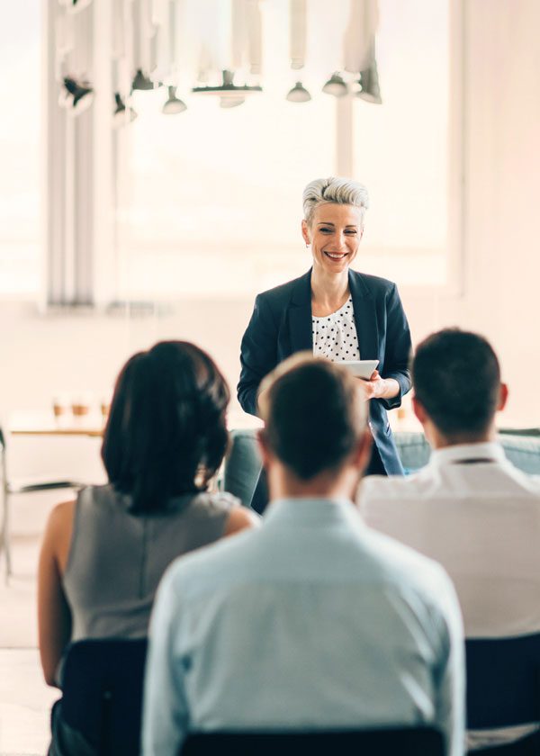Woman with short hair and a black jacket addresses a group of people with their backs to the camera