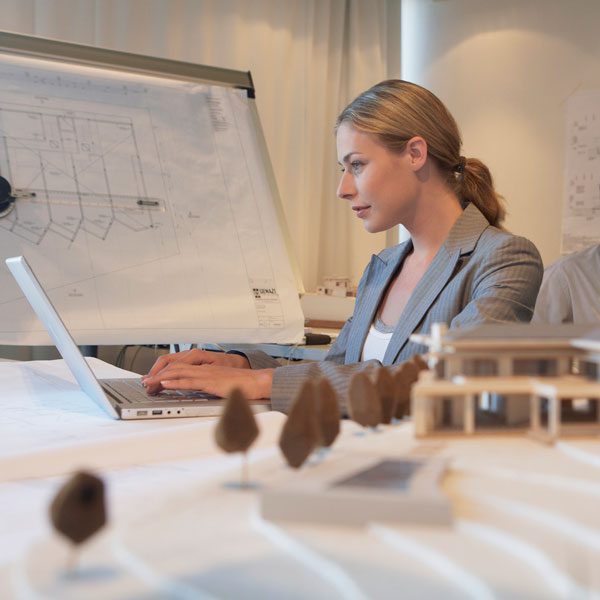 A businesswoman uses a laptop at a table next to a building model slightly blurred in the foreground
