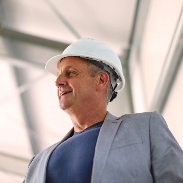Shot from below, an older man in a gray blazer wears a white hard hat