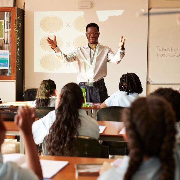 A male teacher stands in front of a projection screen and addresses a classroom full of children