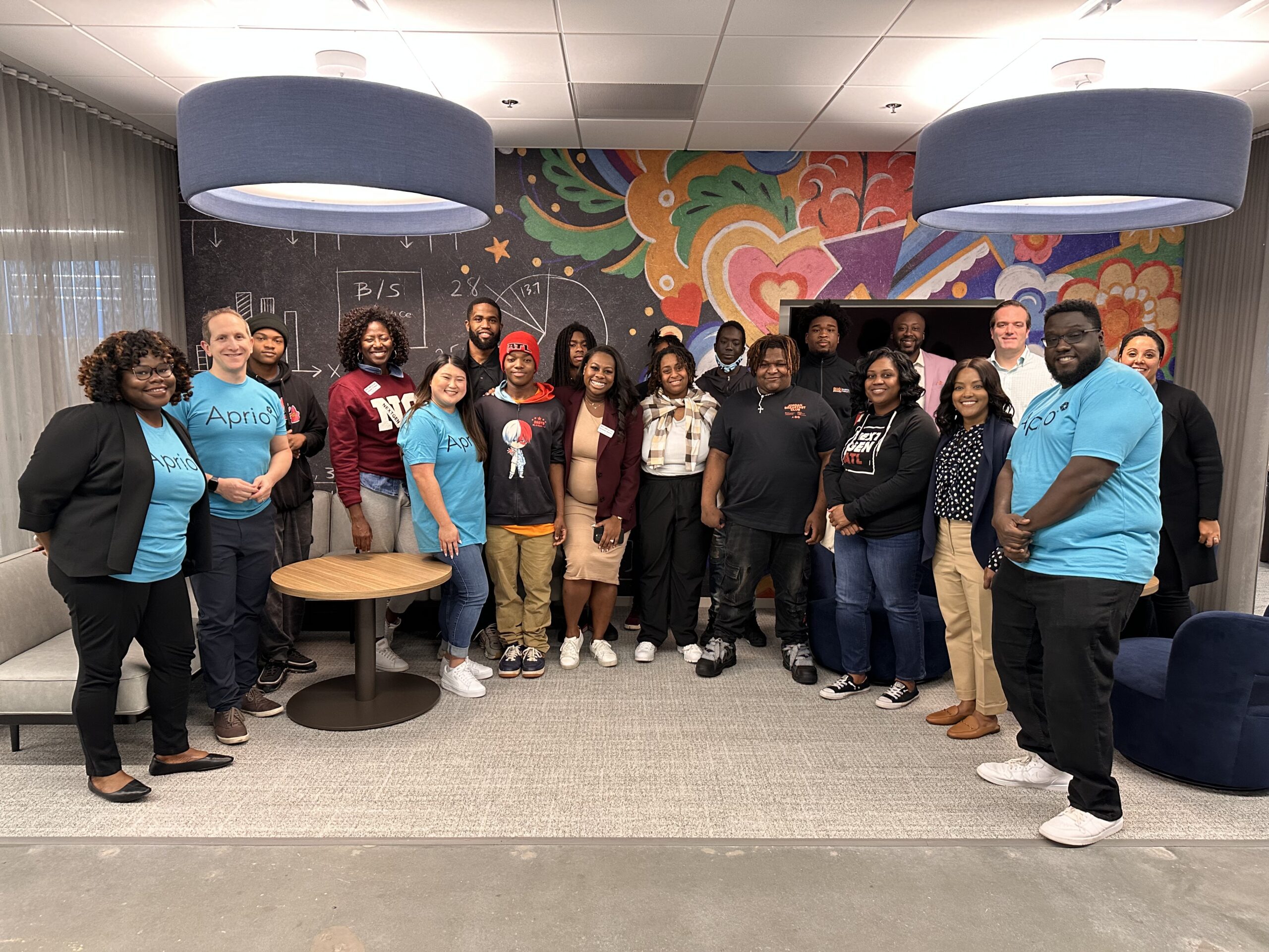 A group of people pose together indoors in front of a colorful mural, some wearing matching blue shirts labeled April.