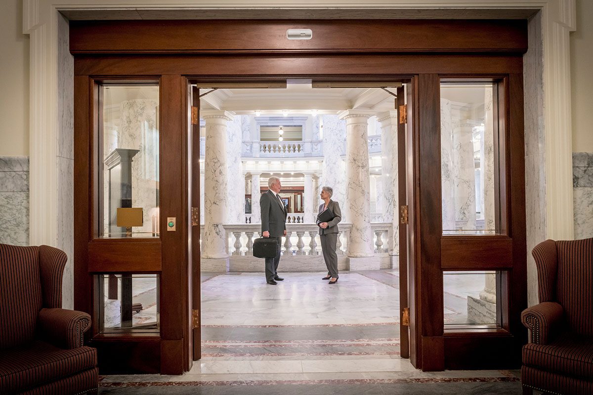 Beyond a handsome wooden double door in a marble building, a man and woman stand amid marble columns