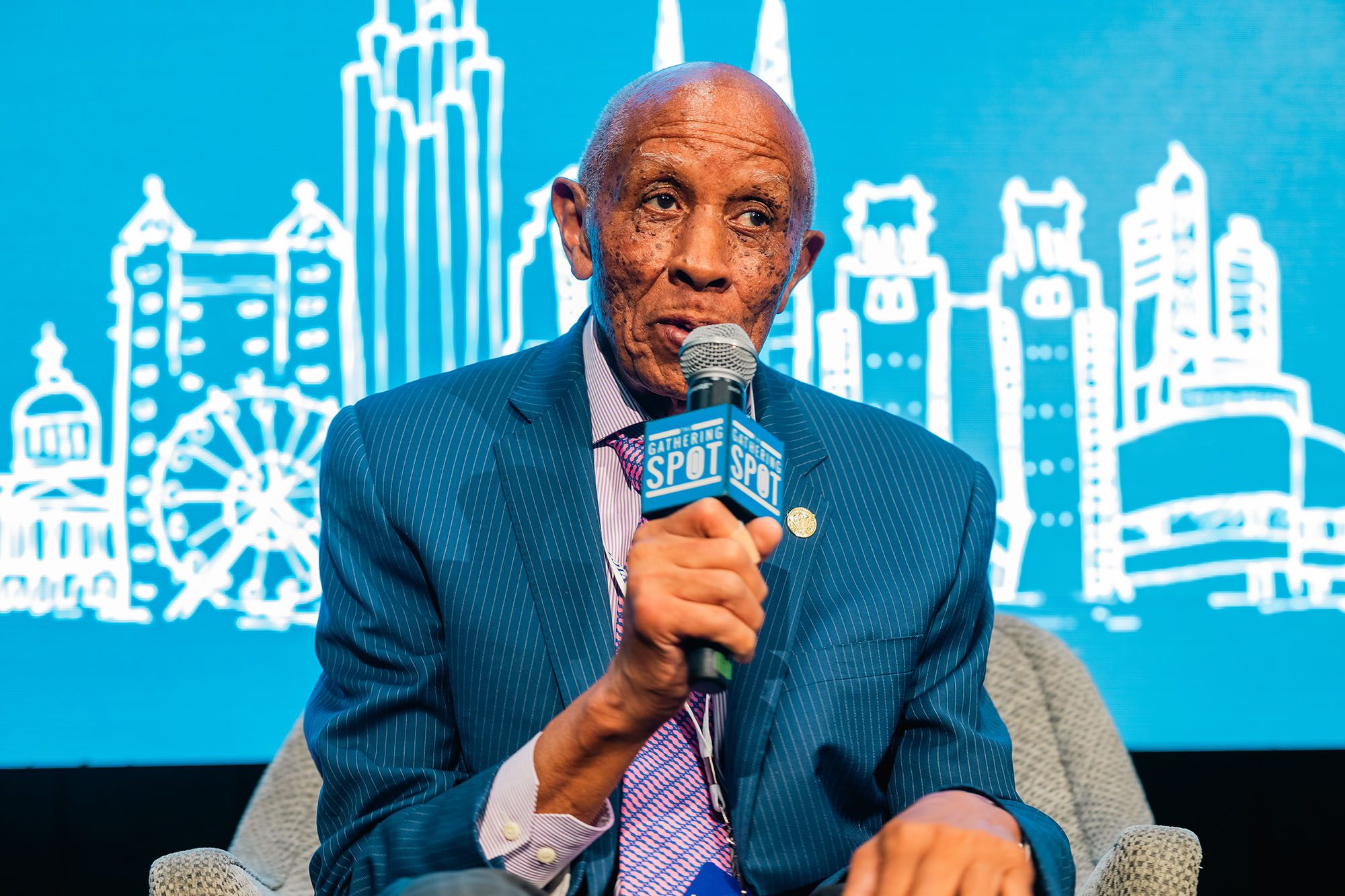 Older man in a blue striped blazer holds a microphone in front of a bright blue screen with white line drawings on it