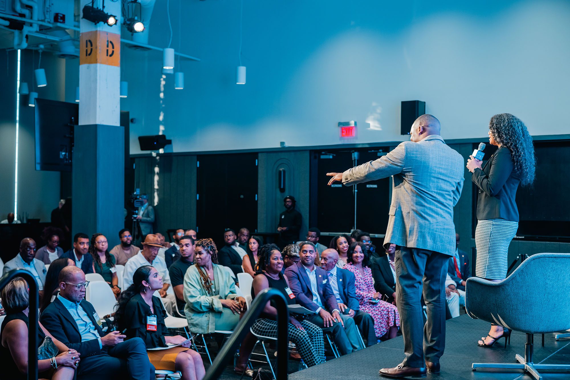 A man and woman stand on a stage with their backs to the camera addressing a crowded event space
