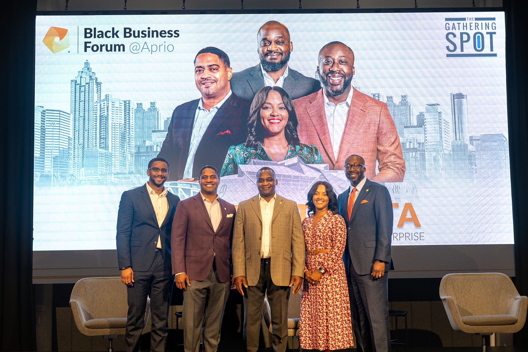 Five people stand on stage in front of a large screen displaying the Black Business Forum @Aprio logo and a group photo with a cityscape background.