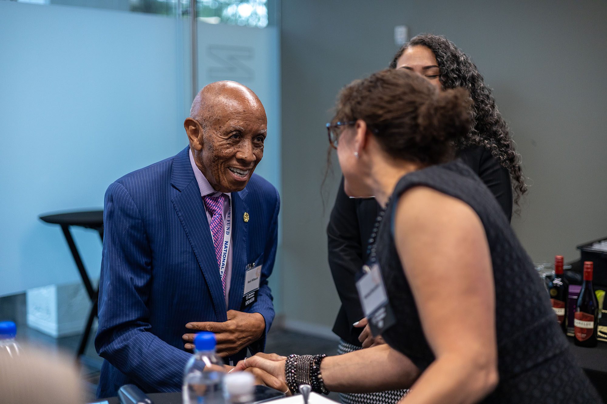 Older man in a blue striped blazer shakes hands with a woman who is facing away from the camera